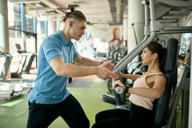istock Young athletic woman using exercise machine with help of her personal trainer in a gym. 1322322479