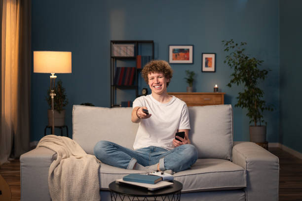 a teenager, a brunette with curly hair relaxes on the living room couch in the evening. the young boy holds the remote control in his hand and switches the channel to a funny program - one person people boredom isolated imagens e fotografias de stock