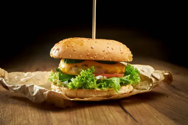 Appetizing burger with chicken, lettuce, melted cheese, cucumber and tomato, served on paper, on a wooden background. Chickenburger