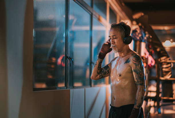 asiático chinês ativo homem boxeador homem de idade fechado ouvindo música com fone de ouvido ao lado janela na academia à noite - muay thai audio - fotografias e filmes do acervo