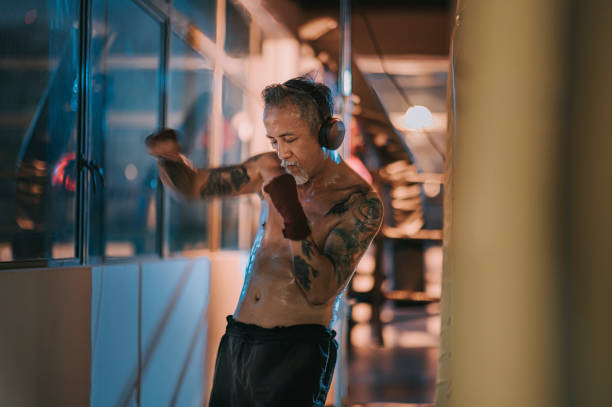 азиатский китайский активный старший боксер глаза человека закрыты практикующих бокс ногами слушать музыку с наушниками рядом с окном в т� - muay thai audio стоковые фото и изображения