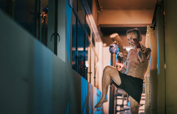 asiático chinês ativo homem boxeador sênior olhos fechado praticando boxe chutando ouvir música com fone de ouvido ao lado janela na academia à noite - muay thai audio - fotografias e filmes do acervo