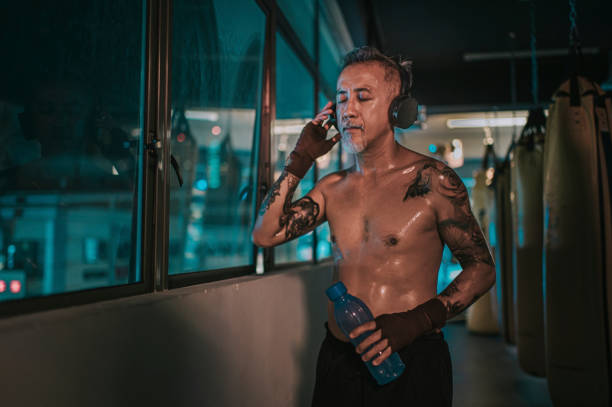 азиатский китайский активный старший боксер глаза человека закрыты слушать музыку с наушниками рядом с окном в тренажерном зале в ночное в - muay thai audio стоковые фото и изображения