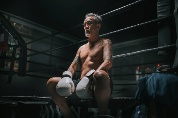 портрет азиатских китайских активных старший боксер человек сидит глядя в сторону рядом с боксерским рингом исчерпаны принимая перерыв - boxing macho men human muscle стоковые фото и изображения