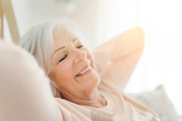 indoor portrait of attractive happy 70 year old senior woman with pleasant smile relaxing on grey sofa in her modern apartment - 60 70 year old imagens e fotografias de stock