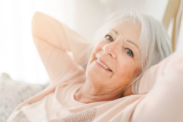 indoor portrait of attractive happy 70 year old senior woman with pleasant smile relaxing on grey sofa in her modern apartment - 60 70 year old imagens e fotografias de stock