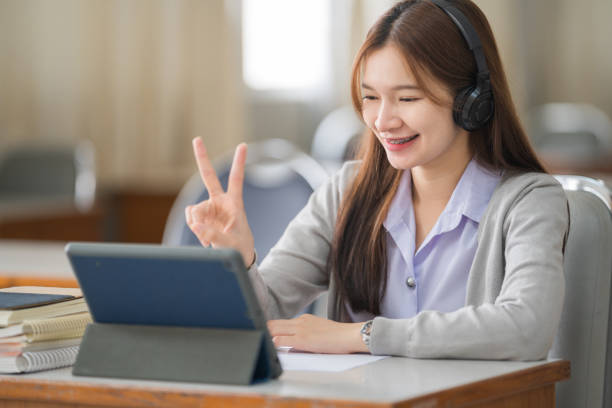 Young interactive happy Asian teenage girl university student studying and presenting her lesson online via video call on a digital tablet in the classroom alone herself. Education stock photo Young interactive happy Asian teenage girl university student studying and presenting her lesson online via video call on a digital tablet in the classroom alone herself. Education stock photo online tutors stock pictures, royalty-free photos & images