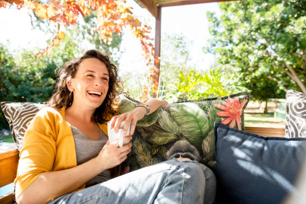 Laughing young woman relaxing with coffee outside on her patio sofa Young woman laughing while relaxing outside on a sofa on her patio and drinking a cup of coffe early fall stock pictures, royalty-free photos & images