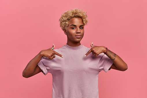 Self assured proud African American woman has blonde hairstyle satisfied with her own high achievements, points with two hands at herself, or at a t-shirt for printing, isolated over pink background.
