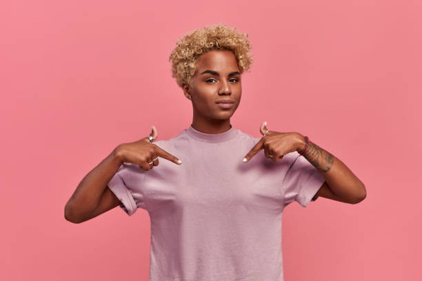 Self assured proud African American woman has blonde hairstyle satisfied with her own high achievements, points with two hands at herself, or at a t-shirt for printing, isolated over pink background Self assured proud African American woman has blonde hairstyle satisfied with her own high achievements, points with two hands at herself, or at a t-shirt for printing, isolated over pink background. arrogance stock pictures, royalty-free photos & images