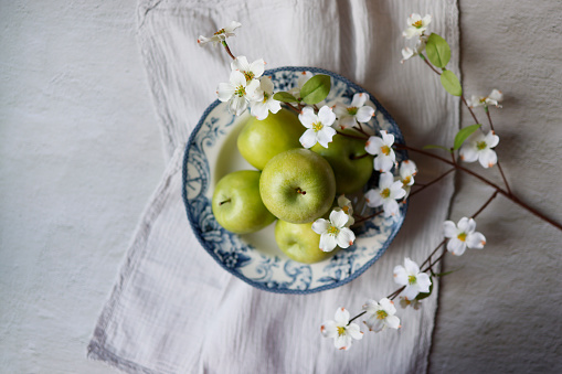Istanbul, Turkey-June 7, 2021: Green apples in a ceramic plate with a blue floral pattern on a light gray rough concrete floor. The plate is on a light gray linen napkin with lace. Around the plate are dogwood branches with white flowers. Full frame, still life, sunlight. Shot with Canon EOS R5.