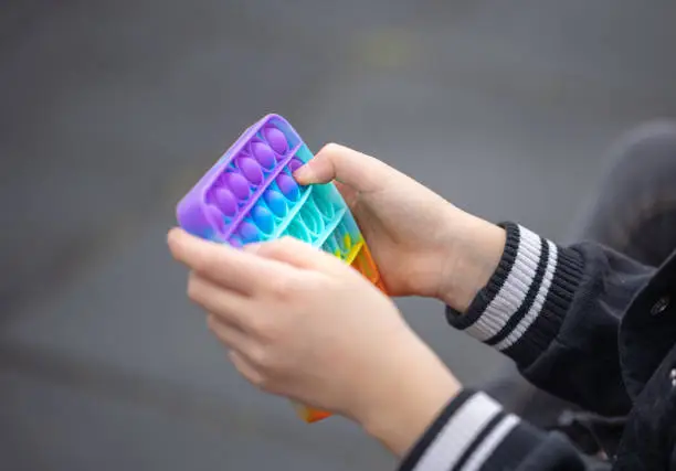 Photo of Close up of kid hands playing with colorful pop It fidget toy.