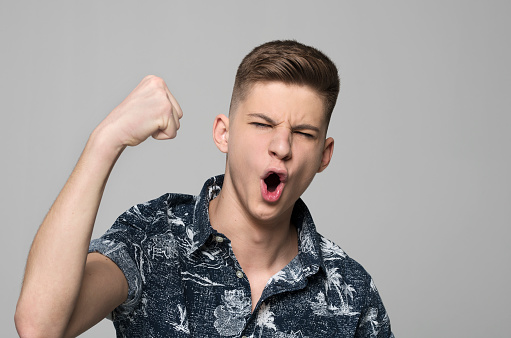 Portrait of excited teenage boy clenching fist while standing against grey background.