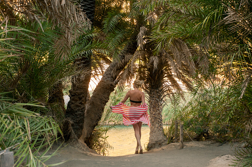 Beautiful happy woman is posing by branches of palm trees at tropical coast in Preveli beach on Crete island.