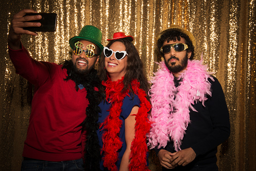 Group of cheerful male and female friends wearing party props while taking selfie with smart phone at photo booth during the party.