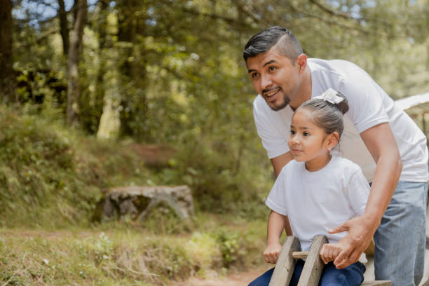 padre con su hija pequeña hispana jugando en el balancín en el parque-niña divirtiéndose su padre en el parque natural - baby mother summer park fotografías e imágenes de stock