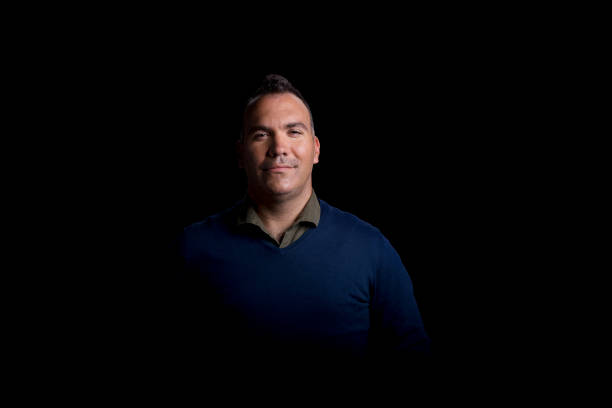 Portrait of smiling young man wearing blue jumper standing against black background.