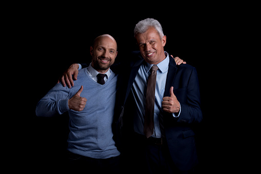 Portrait of smiling male businessmen showing thumbs up while standing against black background.