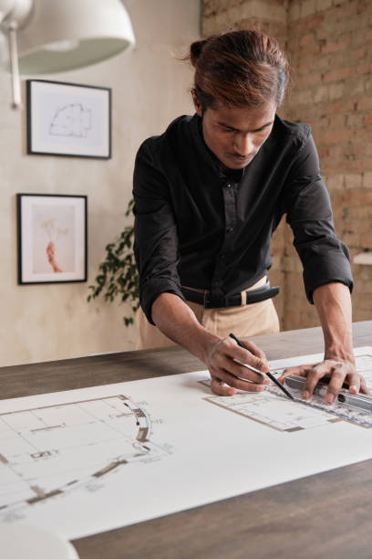 istock Busy young Indian man in black shirt standing at table and using ruler while drafting house plan in loft office with minimalistic pictures on wall 1322370613
