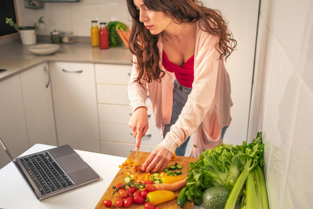 istock Young woman reading online recipes using laptop while cutting vegetables in the kitchen 1322370683
