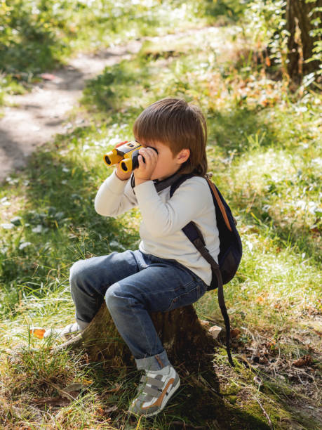 istock Little explorer on hike in forest. Boy with binoculars sits on stump. Outdoor leisure activity for children. Summer journey for young tourist. 1322380079