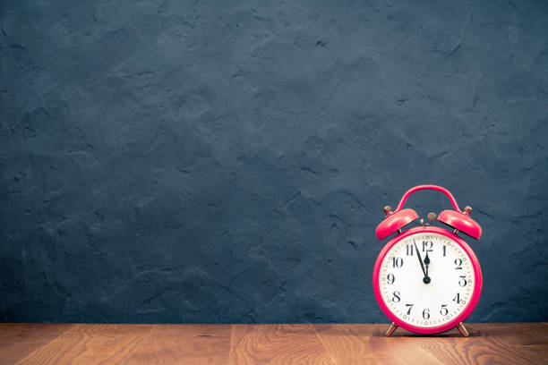 istock Retro alarm clock with five minutes to twelve o'clock on wooden table front old concrete black wall background. Vintage style filtered photo 1322404192