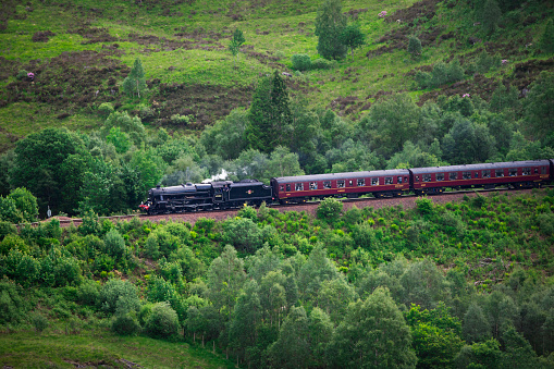 Glenfinnan, UK - June 12, 2019: Passengers travel on a train through forested hills heading toward the Glenfinnan Viaduct Bridge in Scotland