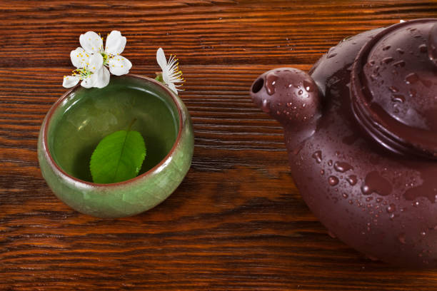 Cup of green tea and clay teapot with cherry flowers on a wooden background.