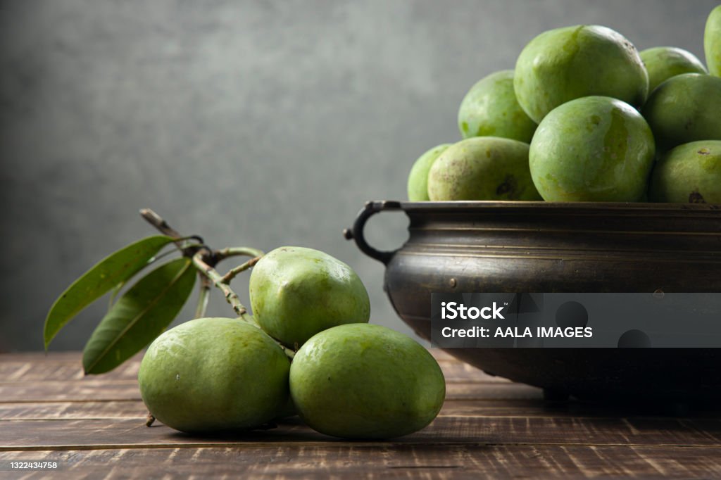 Bunch of Raw mango or Green mango Raw Mango  arranged  traditionally in a brass vessel  and bunch of green mango placed nearby on a wooden textured base with grey background, selective focus. Mango Fruit Stock Photo