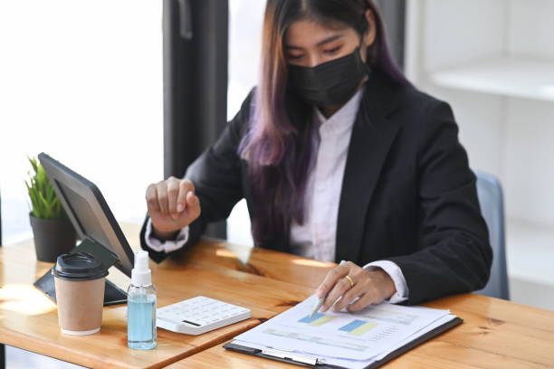 istock Businesswoman in protective mask working with digital tablet and analyzing financial data on wooden desk. 1322442466