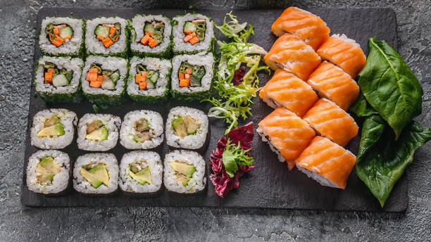 istock Food banner: set of different rolls on a black stone cutting board. Rolls with salmon, vegetables sushi maki rolls and fresh salad. Soy and peanut sauce, chopsticks. Gray concrete background. Top view 1322445473