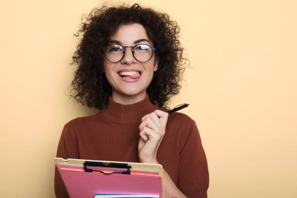 istock Adorable student with curly hair showing her tongue while holding folder on a yellow studio wall 1322450210