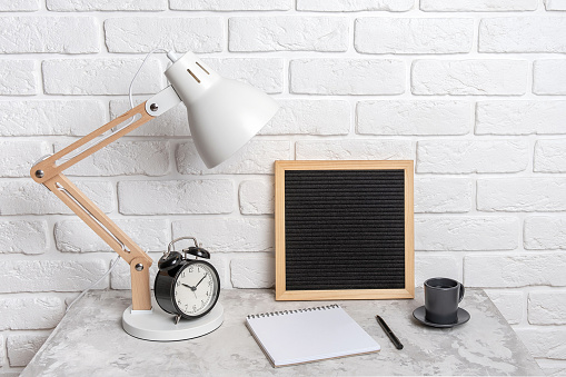 Table lamp, empty blank letter board, notebook and alarm clock on the table, against the background of a white brick wall. Workplace, front view, mock up.