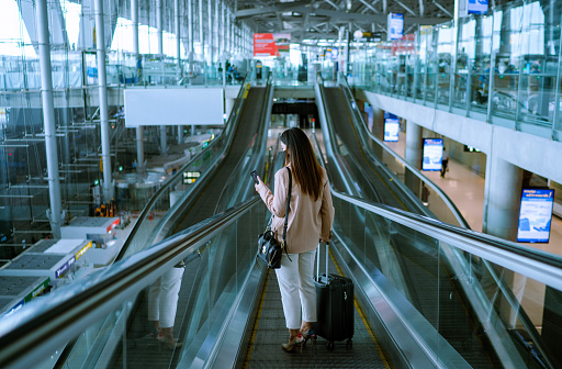 Asian businesswoman traveler with face mask on the move using escalator in airport.