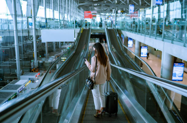 azjatycka bizneswoman podróżniczka z maską na twarzy w ruchu za pomocą schodów ruchomych na lotnisku. - arrival departure board airport travel business travel zdjęcia i obrazy z banku zdjęć