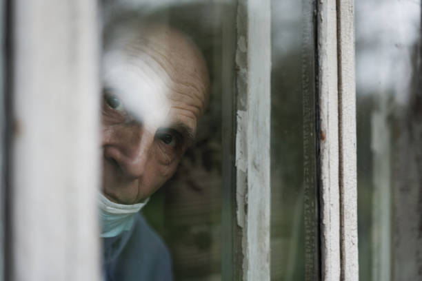 Old solitary man wearing face mask looking through window.