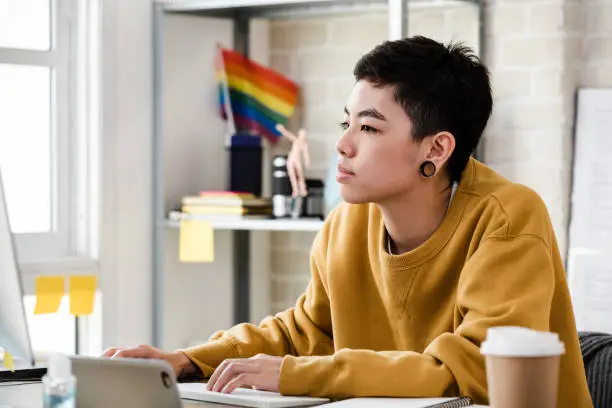 Photo of Young Asian tomboy woman in casual attire working from home on laptop computer in living room