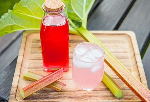 Homemade rhubarb syrup ( Rheum rhabarbarum ). Nice pink liquid syrup in bottle and glass with juice and ice cubes in drinking glass on tray, decorated with rhubarb stalks. Refreshing spring drink.