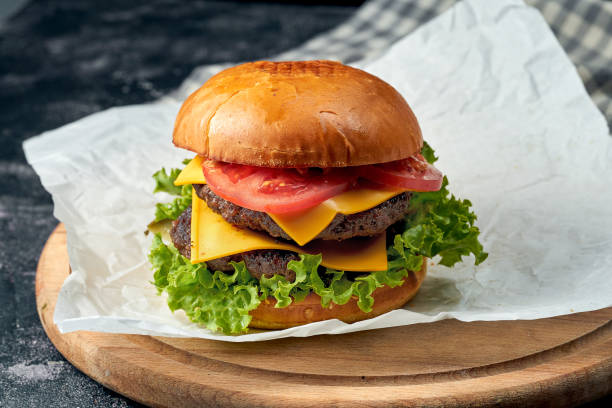 istock Delicious double burger with beef, tomatoes, lettuce, melted cheese on white parchment. Dark background. American fast food 1322469922
