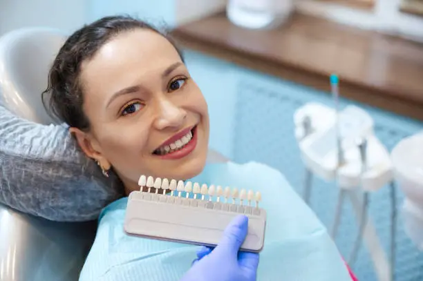 Brunette woman with beautiful smile before receiving dental care check up and teeth whitening bleaching, female doctor wearing exam gloves checks tooth color with a comparison veneer scale chart.