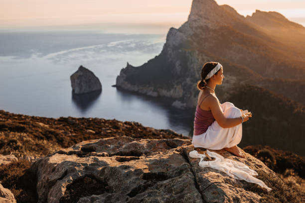 Young woman sitting with her little diary book in the mountains while doing meditation during the sunrise in front of an amazing seascape stock photo
