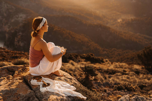 Beautiful girl sitting with her little diary book in the mountains while doing meditation during the sunrise in front of an amazing landscape stock photo