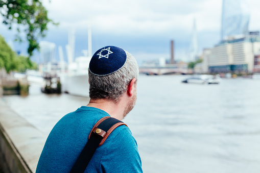 Color image depicting a mid adult Jewish man in his 30s wearing a traditional Jewish skull cap (with star of David design). He is on a city street in central London.