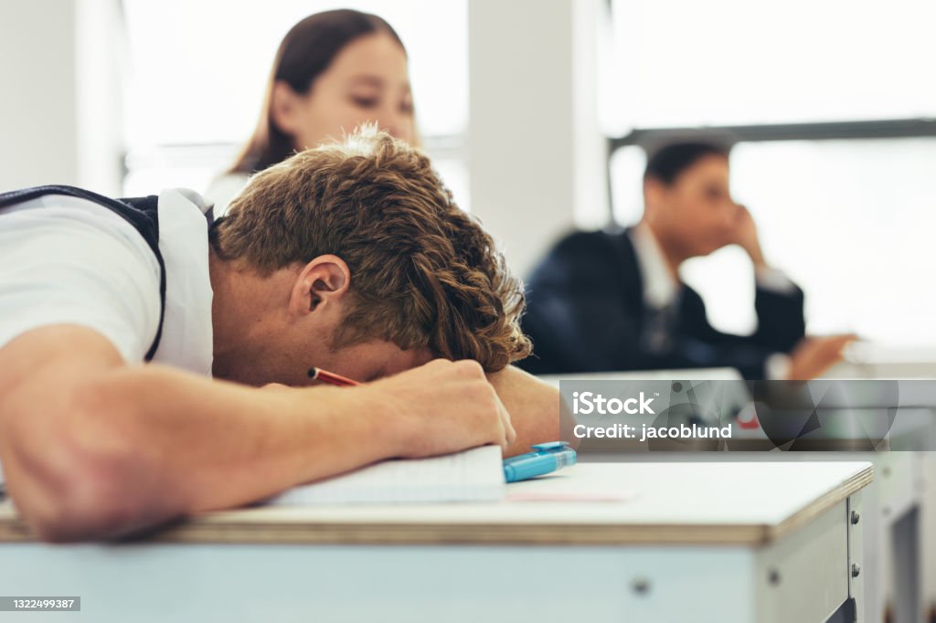 Bored high school student sleeping on classroom desk Bored male high school student sleeping during the lecture in the classroom. Schoolboy sleeping on desk in classroom with classmates in background. Classroom Stock Photo