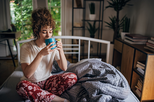 One woman, young female in pajamas sitting in her bed at home. She is enjoying in morning coffee.