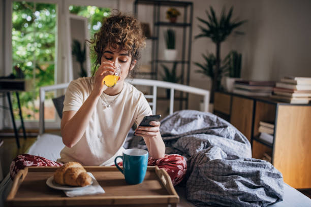 Beautiful morning in bed One woman, young female in pajamas sitting in her bed at home. She is having breakfast in bed and using smart phone. juice drink stock pictures, royalty-free photos & images