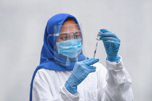 Close-up shot of muslim Indonesian healthcare worker filling a syringe with the Covid-19 vaccine at hospital. She's wearing a blue hijab, face shield, surgical mask and surgical gloves for safety protection.