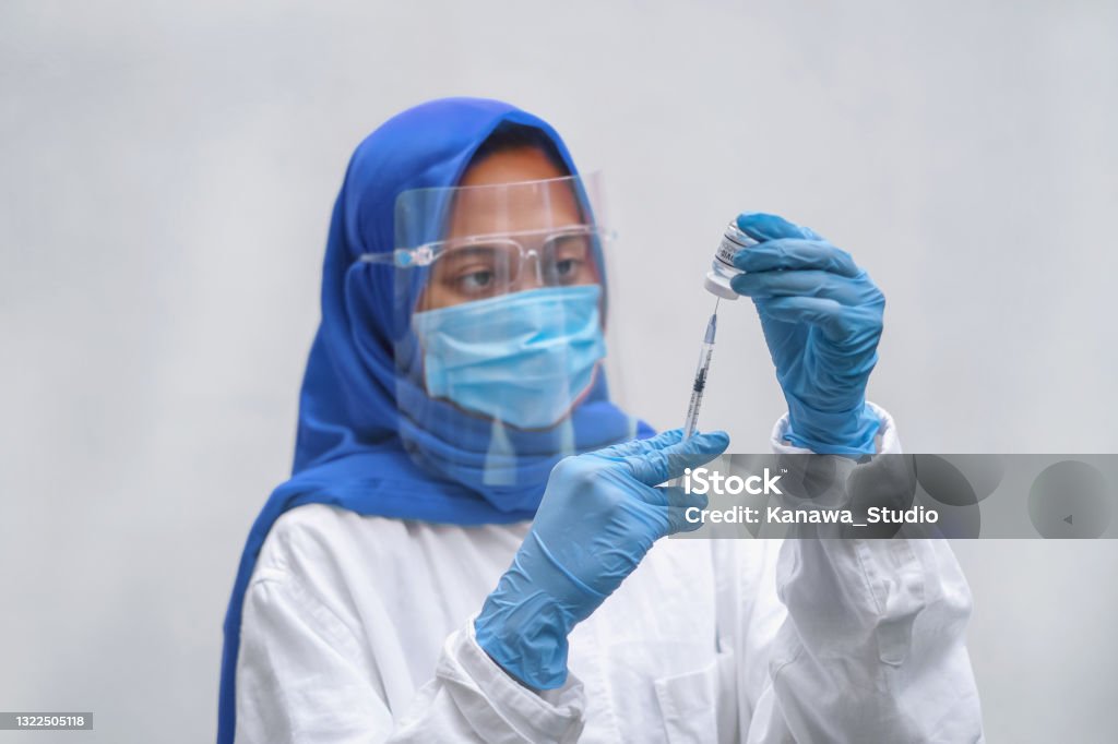 Indonesian Healthcare Worker Filling a Syringe With the Vaccine Close-up shot of muslim Indonesian healthcare worker filling a syringe with the Covid-19 vaccine at hospital. She's wearing a blue hijab, face shield, surgical mask and surgical gloves for safety protection. Nurse Stock Photo