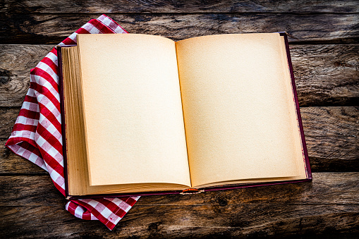 Open vintage cookbook with blank pages shot from above on rustic wooden table. A checkered tablecloth is under the cookbook. Useful copy space available for text and/or logo on book pages.