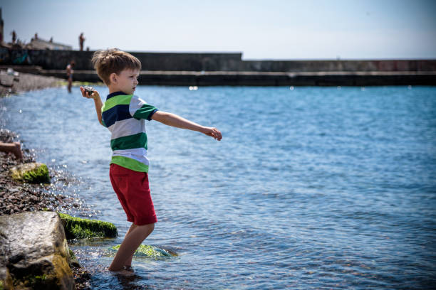 o menino à beira-mar joga pedras na água. relaxamento e férias com crianças. infância feliz - throwing stone little boys child - fotografias e filmes do acervo